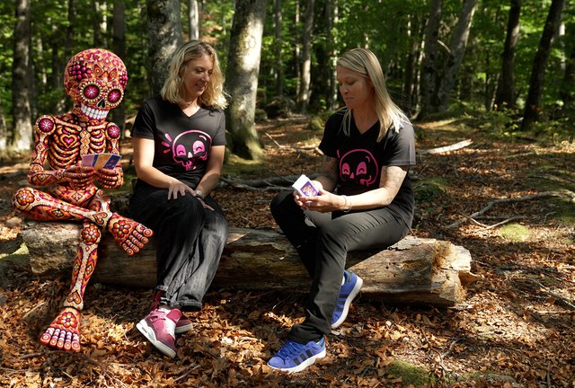 Le t-shirt De la mort qui tue porté en forêt Deux personnes assises en forêt, portant le t-shirt De la mort qui tue à crâne rose, avec la figurine squelette de la marque